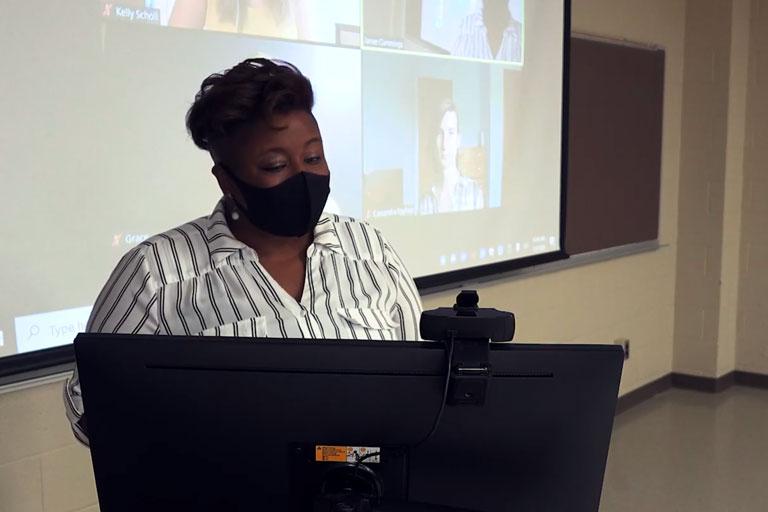 Photo of an instructor at the teaching computer with students participating via Zoom behind her
