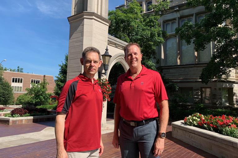 Greg Siering and Randy Newbrough, both in red shirts, standing at Indiana University's Sample Gates