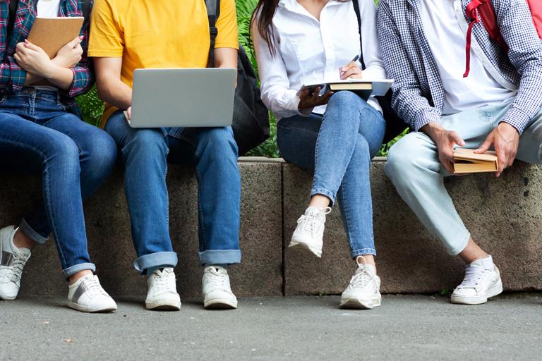 Four individuals shown from chest down, sitting on a stone wall, holding various books, notebooks, and computers
