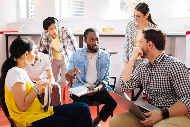 Five students are gathered around and listening to a sixth student in a classroom setting.