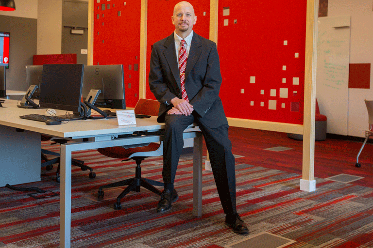 James McGookey sitting on the edge of a desk, looking at the camera, wearing a suit. There is a red room divider in the background. 