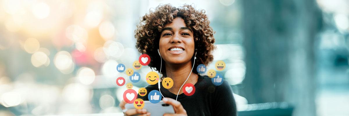 A young woman with earbuds in and emojis floating in the air around her device